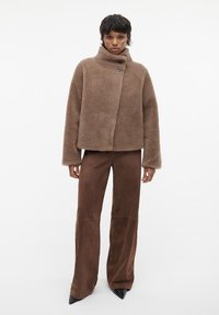 Bruine donzige wollen jas met een hoge kraag en knoopsluiting, gecombineerd met bruine suede wijde broek en zwarte puntige schoenen.