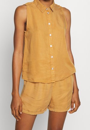 Bezrękawnik zapinany na guziki w kolorze musztardowym, zestawiony z luźnymi szortami w tym samym kolorze, noszony przez osobę z ramionami swobodnie opuszczonymi wzdłuż ciała.