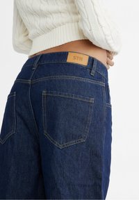 Dark blue denim jeans featuring back pockets, a textured surface, and a tan leather tag with "STR" at the waistband.