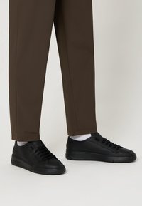 Baskets en cuir noir avec une surface texturée et des semelles en caoutchouc noir, associées à un pantalon droit marron et des chaussettes blanches.