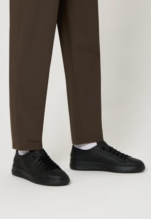 Sneakers in pelle nera con superficie testurizzata e suole in gomma nera, abbinate a pantaloni dritti marroni e calzini bianchi.