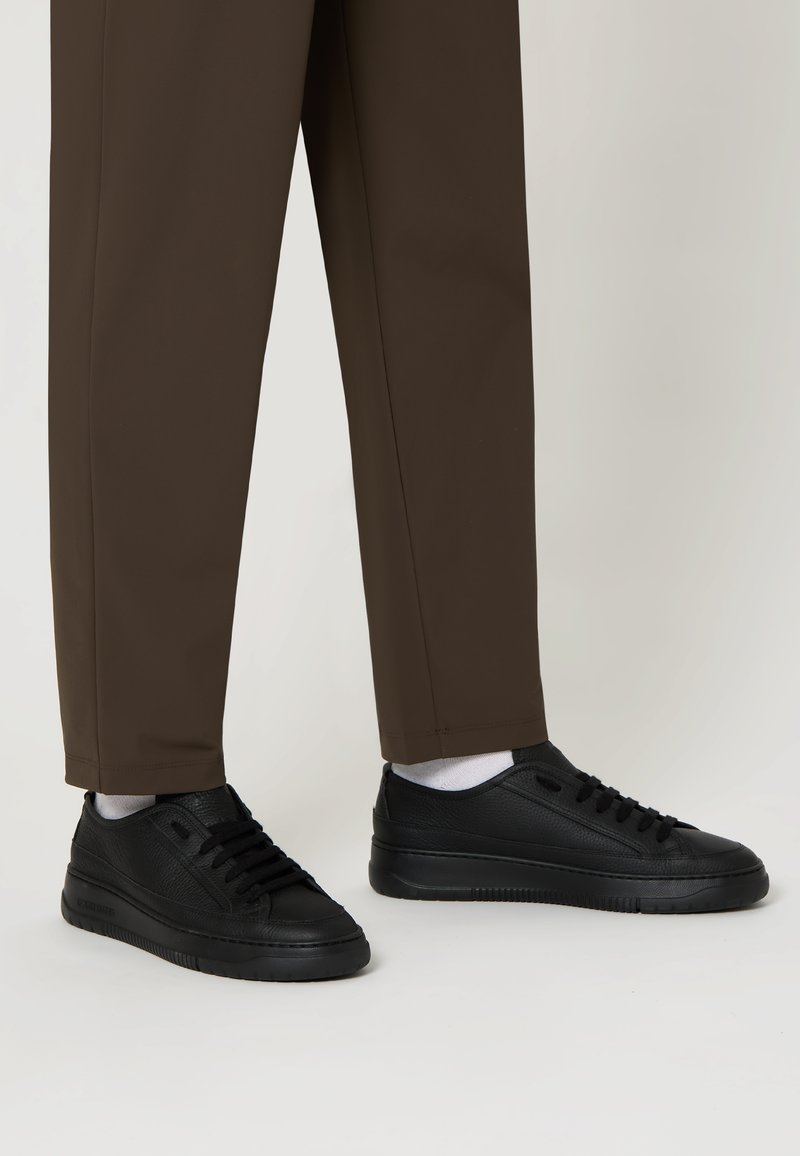 Baskets en cuir noir avec une surface texturée et des semelles en caoutchouc noir, associées à un pantalon droit marron et des chaussettes blanches.