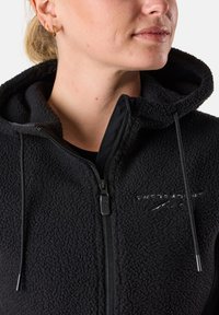 Schwarze Fleece-Jacke mit Kapuze, ausgestattet mit einem Reißverschluss, Kordelzügen und gesticktem Logo. Weiche Textur mit einem leicht plüschigen Aussehen.