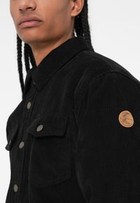 Schwarze Cordjacke mit Kragen, ausgestattet mit zwei Brusttaschen, Metall-Druckknöpfen und einem runden Lederlogo-Patch am Arm.