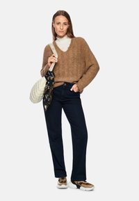 Brauner, flauschiger Pullover mit V-Ausschnitt, dunkle Jeans und gemusterte Turnschuhe. Hält eine gesteppten cremefarbigen Tasche mit einem schwarzen Schalakzent.