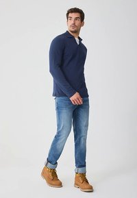 Laivastonsininen ribbineulottu pitkähihaisen poolopaidan, vaaleansiniset farkut ja vaaleanruskeat kengät kultaisilla yksityiskohdilla. Malli kävelee valkoista taustaa vasten.