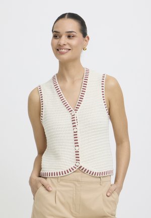 Kvinde smilende, iført en hvid strikket ærmeløs vest med brune stribede kanter og beige bukser, hænder i lommerne, foran en ensfarvet baggrund.