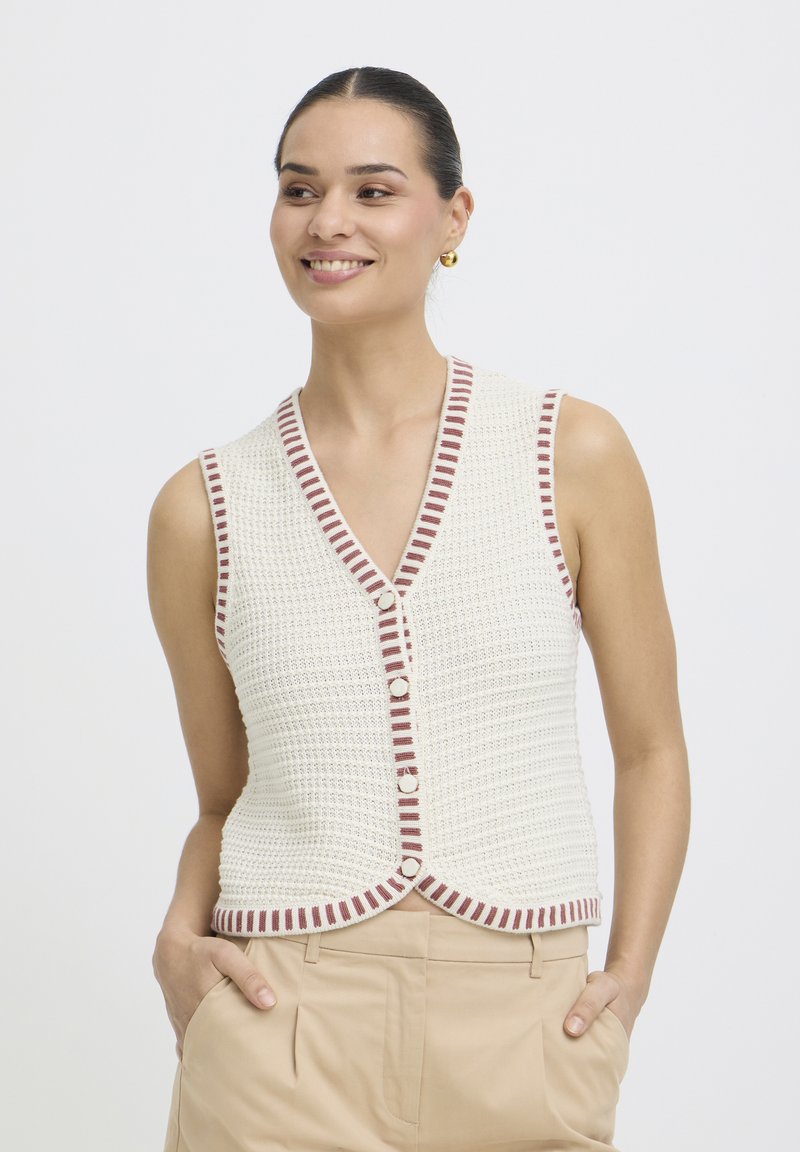 Naeratav naine, seljas valge heegeldatud varrukateta vest, millel on pruunid triibulised ääred, ning beežid püksid, käed taskutes, ühevärvilise taustaga.