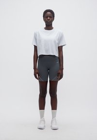 Ψηλός άτομο που στέκεται μπροστά, φοράει λευκό κοντό μπλουζάκι, σκούρα γκρι ποδηλατικά σορτς, λευκές κάλτσες και λευκά αθλητικά παπούτσια σε απλό φόντο.