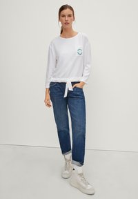 Maglietta bianca a maniche lunghe con vita annodata, caratterizzata da una tasca ricamata con logo in teal. Abbinata a jeans blu a fit rilassato e sneakers bianche.