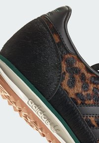 Mustikkapisteinen musta ja ruskea leopardi-kuosisneaker, jossa on poniturkista, sileitä nahkaosia ja teksturoitu kumipohja vihreällä reunuksella sekä Adidas-logolla.