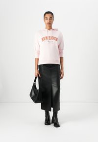 Felpa rosa con la scritta "NEW HAVEN", abbinata a una gonna midi in finta pelle nera con uno spacco frontale. Tiene in mano una borsa nera e indossa stivali neri.