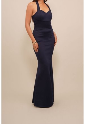 Vrouw die een mouwloze, marineblauwe galajurk in zeemeerminstijl met halternek draagt, staand tegen een effen beige achtergrond.