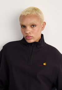 Zwarte kwart-rits sweatshirt met een zachte textuur, voorzien van een klein geborduurd Carhartt-logo in geel en rood op de borst.