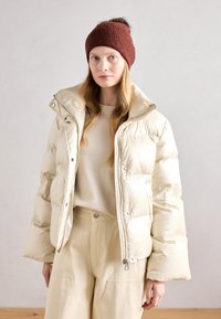 Chaqueta acolchada de color beige claro con cuello alto, acabado brillante, cierre de cremallera y acentos de botón. Lleva encima un suéter crema y pantalones beige.