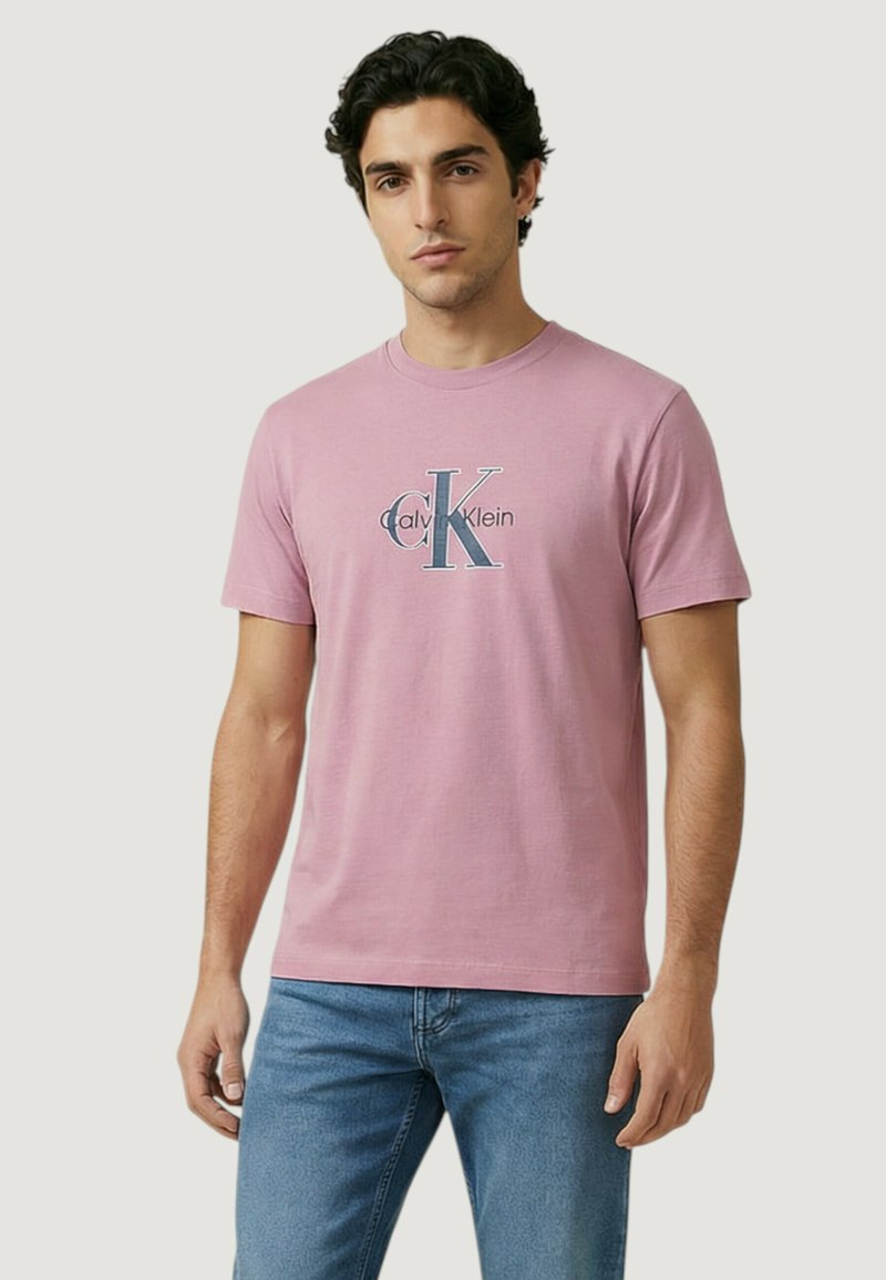 Jeune homme portant un t-shirt rose avec le logo Calvin Klein et un jean bleu, debout devant un fond clair uni.