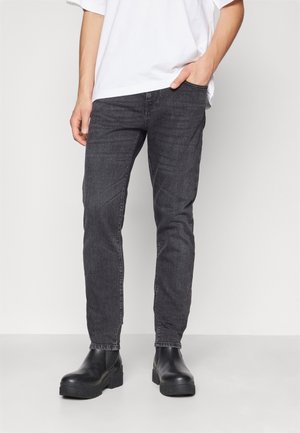 Person trägt dunkelgraue Slim-Fit-Jeans, schwarze klobige Stiefel und ein weißes T-Shirt und steht mit einer Hand in der Tasche.