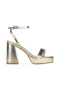 Sandalia de tacón alto de metal dorado con un tacón bloque grueso, correa ajustable en el tobillo y correas delanteras anchas. Textura suave y diseño minimalista.