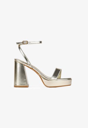 Metallisch goldene High-Heel-Sandale mit einem klobigen Blockabsatz, verstellbarem Knöchelriemen und breiten Vorderbändern. Glatte Textur und minimalistisches Design.