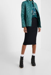 Türkis gesteppte Jacke mit Stehkragen, getragen über einem schwarzen, figurbetonten Kleid. Schwarze Knöchelstiefel mit Reißverschlussdetails und weißen Knöchelsocken runden den Look ab.