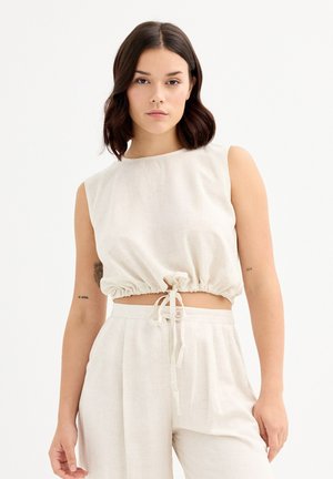 Top corto sin mangas de color beige claro en tela suave, con un diseño fruncido y cordón en la cintura, y un cuello redondo.