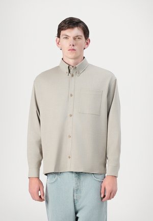 Beige button-up shirt with a collar, long sleeves, a chest pocket, and subtle texture, paired with light blue denim jeans.