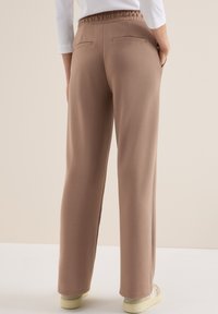 Bruine broeken met elastische tailleband, achterzakken, wijd uitlopende snit, gladde textuur en een relaxte fit, gecombineerd met lichtgekleurde sneakers.