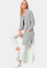 Lichtgrijze cardigan met lange mouwen en zakken, gedragen over een gestreept topje en witte broek, gecombineerd met witte sneakers. Gladde textuur.