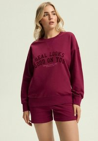 Burgundowy zestaw z bluzą i szortami; materiał bawełniany o luźnym kroju, z czarnym napisem "REAL LOOKS GOOD ON YOU" oraz ściągaczami.