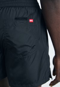Juodi maudymosi šortai su elastingu juosmeniu, turintys užpakalinį kišenę ir raudoną Diesel logotipo ženklelį. Lygus, lengvas audinys su matinio paviršiumi.
