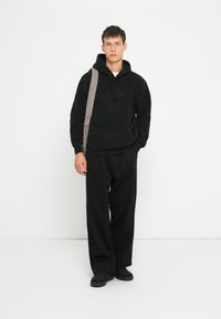 Schwarze Fleece-Hoodie mit Kängurutasche und tonalem Logo, kombiniert mit lockeren schwarzen Hosen und schwarzen Slippern. Grauer Schultergurt.