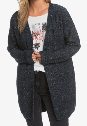 Donkerblauwe gebreide cardigan met een open voorkant, lange mouwen en geribbelde manchetten. Draagt over een grafisch wit T-shirt en zwarte broek.