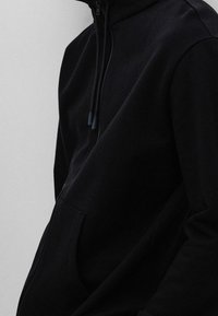 Primer plano de una persona que lleva una sudadera con cremallera negra, con cordones y un bolsillo frontal, contra un fondo liso.