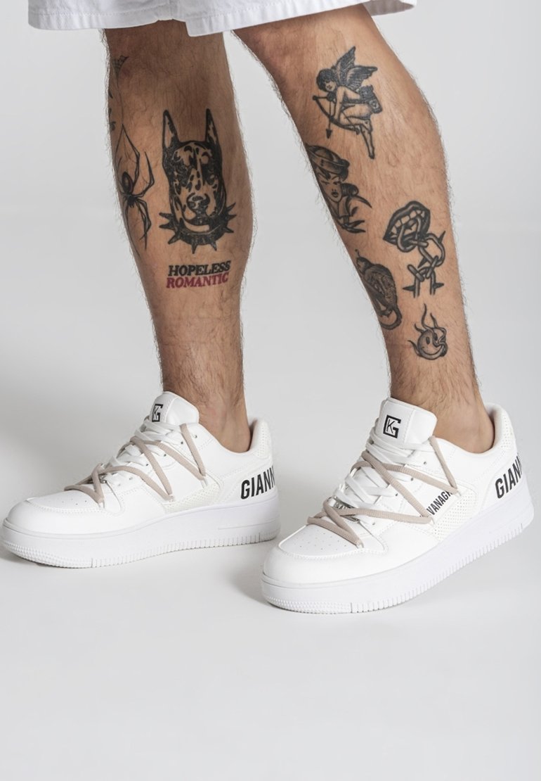 Piernas con tatuajes negros que incluyen un perro, una araña, un cupido y el texto "ROMÁNTICO SIN ESPERANZA"; llevan zapatillas blancas Giannv con cordones beige.