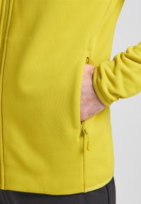 Geel softshell jack met een gladde textuur, voorzien van een zijzak met rits en een aansluitend manchetdetail.