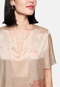 Seidenbluse in sanftem Pfirsich mit floralem Muster, kurzen Ärmeln, V-Ausschnitt und Nadelstreifendetails auf der Vorderseite. Bronzenfarbene runde Ohrringe.