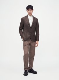 Homme debout portant un blazer marron à carreaux, une chemise blanche boutonnée, un pantalon beige et des chaussures habillées noires sur un fond uni.