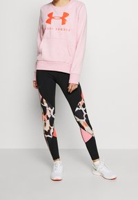 Růžová mikina s výrazným oranžovým logem Under Armour, spárovaná s černými legínami s barevným abstraktním vzorem a růžovými akcenty.
