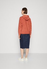 Sudadera con capucha de color óxido, con textura y estampado de logotipos, combinada con una falda de mezclilla oscura y zapatillas blancas, vista desde atrás.