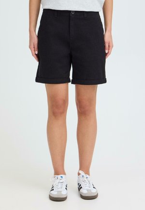 Kvinne iført svarte shorts med oppbrettet kant, hvite joggesko med svarte striper, og en lys topp, stående mot en ensfarget bakgrunn.