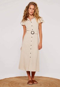 Beiges Leinenkleid mit kurzen Ärmeln, Knopfverschluss und einem runden Gürtelakzent. Modell trägt braune geflochtene Sandalen und steht auf einem geflochtenen Teppich.