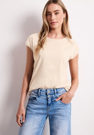Jeune femme aux longs cheveux blonds portant un t-shirt beige et un jean bleu clair, debout avec une main dans la poche devant un fond blanc.