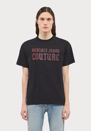 T-shirt di cotone nero con stampa grafica di "VERSACE JEANS COUTURE" in un carattere audace e ornamentale sul petto. Maniche corte.