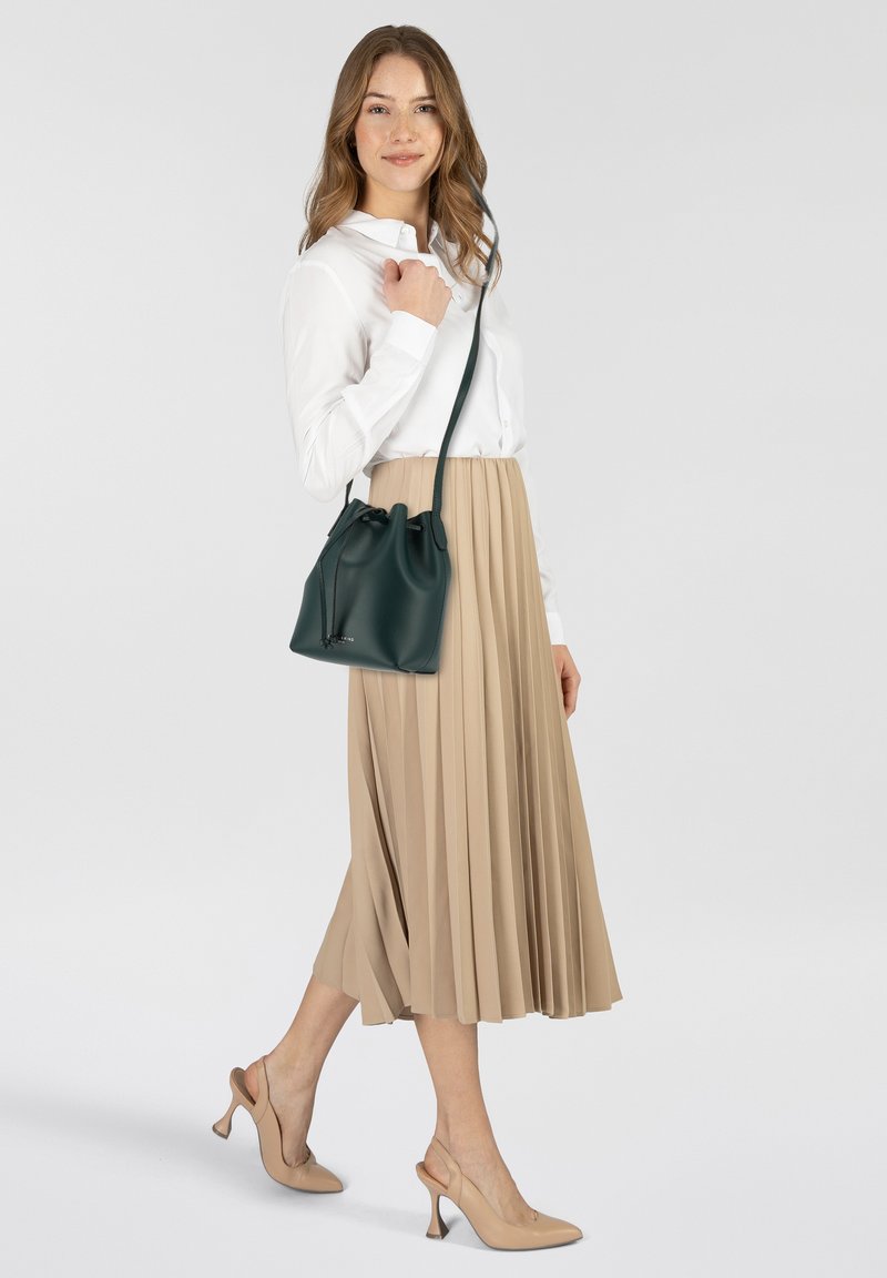Borsa a secchiello verde in pelle con chiusura a corda, superficie testurizzata e tracolla regolabile. Indossata con una gonna beige a pieghe e una camicia bianca.