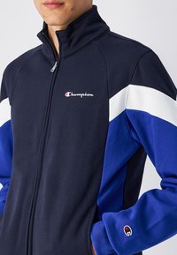 Giacca a zip di tuta di colore blu navy con maniche bloccate in bianco e blu, realizzata in materiale poliestere, e un piccolo logo rosso e bianco sul petto.