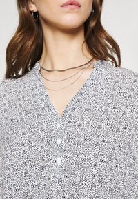 Kobieta w wzorzystej białej bluzce z guzikami, z nałożonymi cienkimi srebrnymi naszyjnikami oraz srebrną kolczykiem w kształcie koła, ukazana od podbródka do klatki piersiowej.