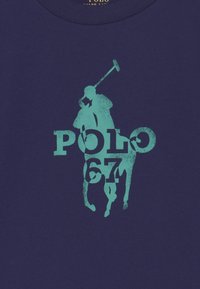 Camiseta de algodón azul marino con un gráfico de un jugador de polo turquesa que incorpora el texto "POLO 67", destacando colores llamativos y un diseño sencillo.