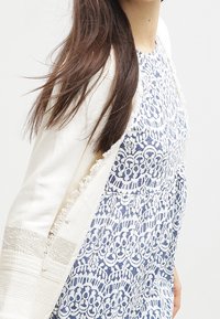 Valkoinen neule, jossa on tekstuuri ja purjeet kiristykset, kerrostettuna sinisen mekon päälle, jossa on monimutkaisia valkoisia pitsikuvioita.