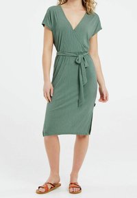 Vestido verde acanalado tipo envolvente con mangas cortas, escote en V profundo, cintura ajustada con lazo y aberturas laterales. Combinado con sandalias marrones con tiras.