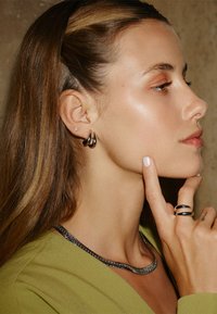 Svart- och silverfärgade örhängen med droppliknande form; ett silverkedjehalsband; ringar med en slät design; modellen har rakt, långt hår.